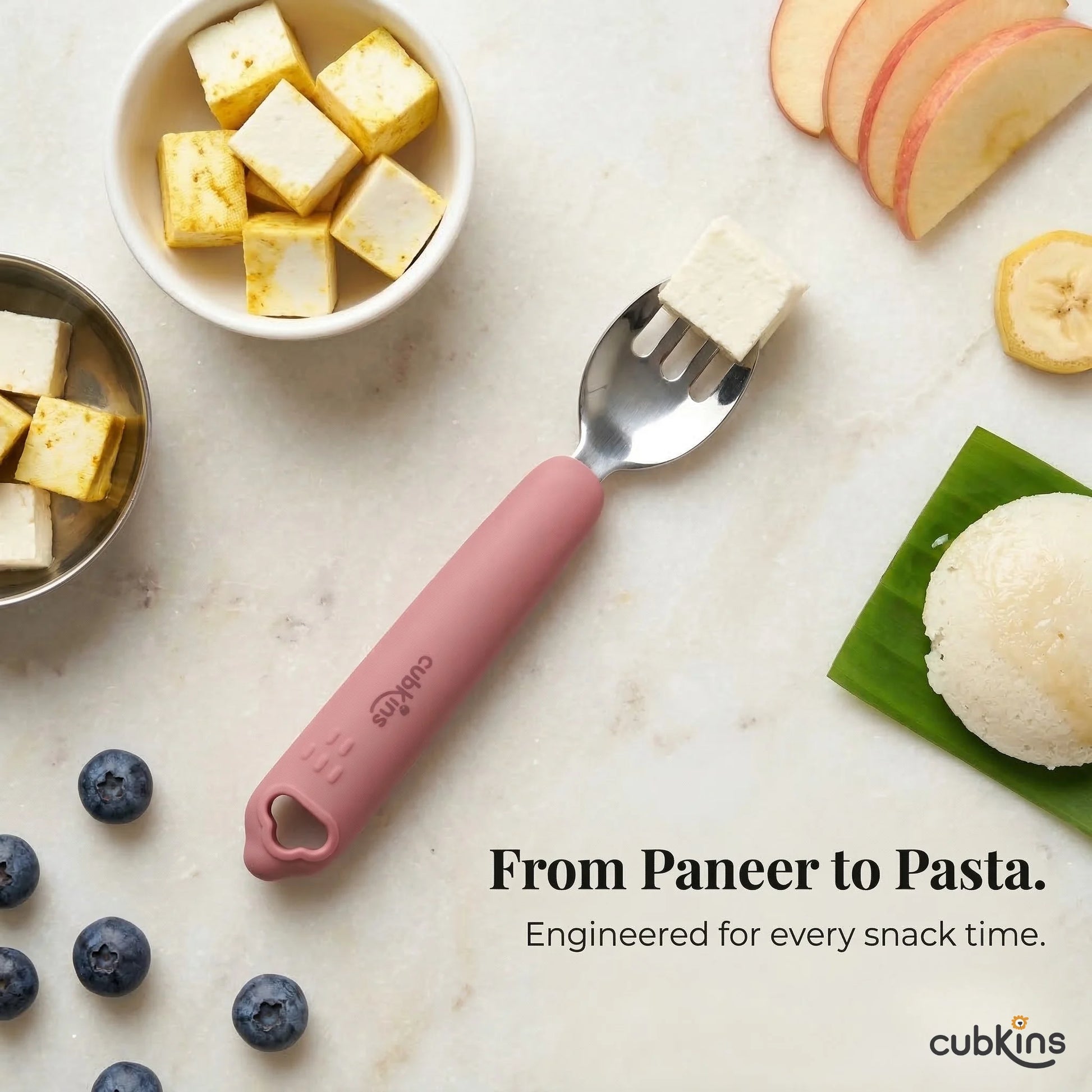 Cubkins Raspberry toddler fork with a cube of paneer on the tines, surrounded by Indian snack foods including idli, paneer, blueberries, and apple slices, with the caption "From Paneer to Pasta. Engineered for every snack time."