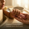 Close-up of a baby and parent's hands holding a Cubkins Raspberry toddler fork together as the baby eats a piece of banana, with the caption "Guided to Independence – Ergonomic grip for shared control"