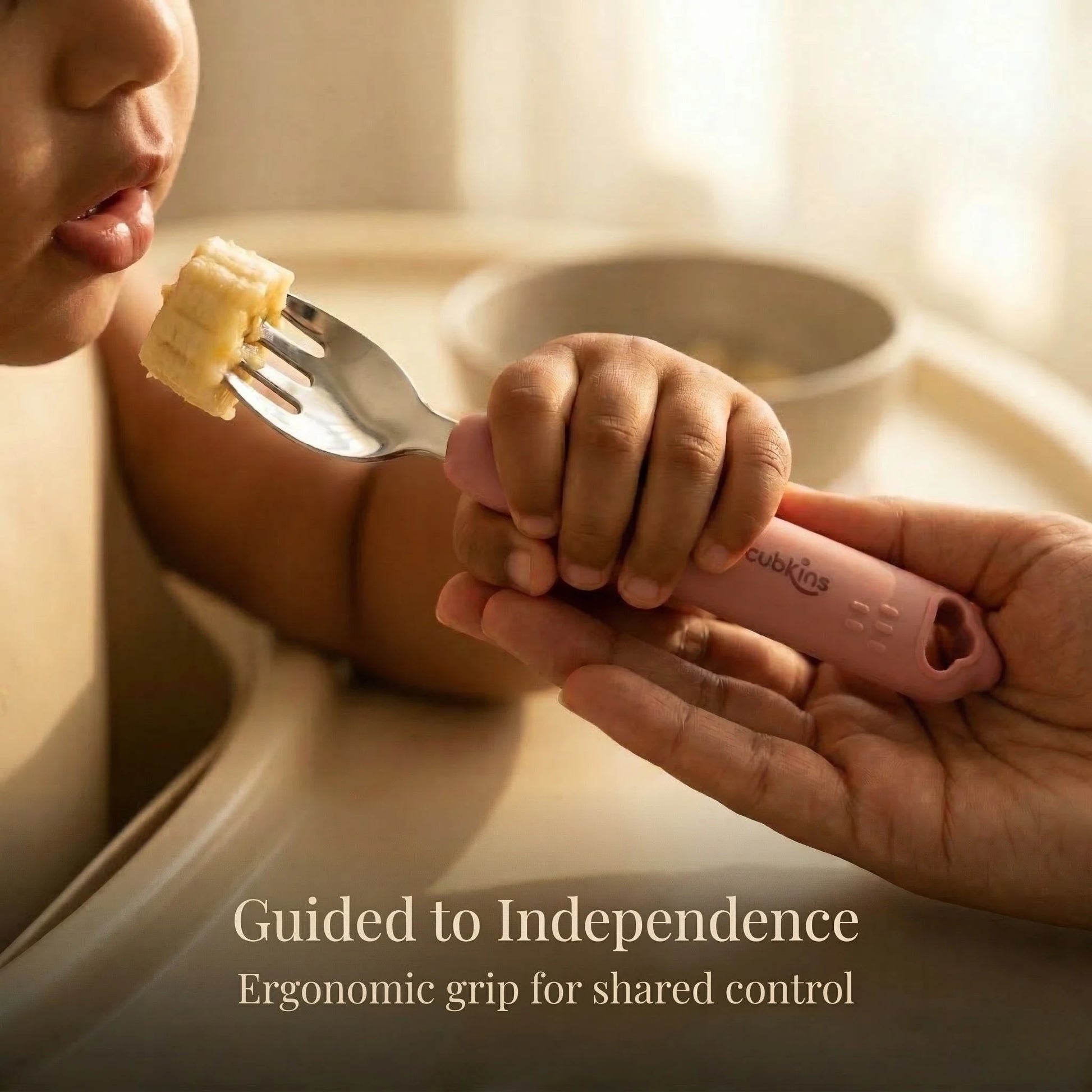Close-up of a baby and parent's hands holding a Cubkins Raspberry toddler fork together as the baby eats a piece of banana, with the caption "Guided to Independence – Ergonomic grip for shared control"