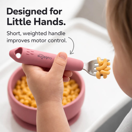 Toddler's hand holding a Cubkins Raspberry fork with macaroni on the tines above a matching raspberry silicone bowl, with the text "Designed for Little Hands – Short, weighted handle improves motor control"