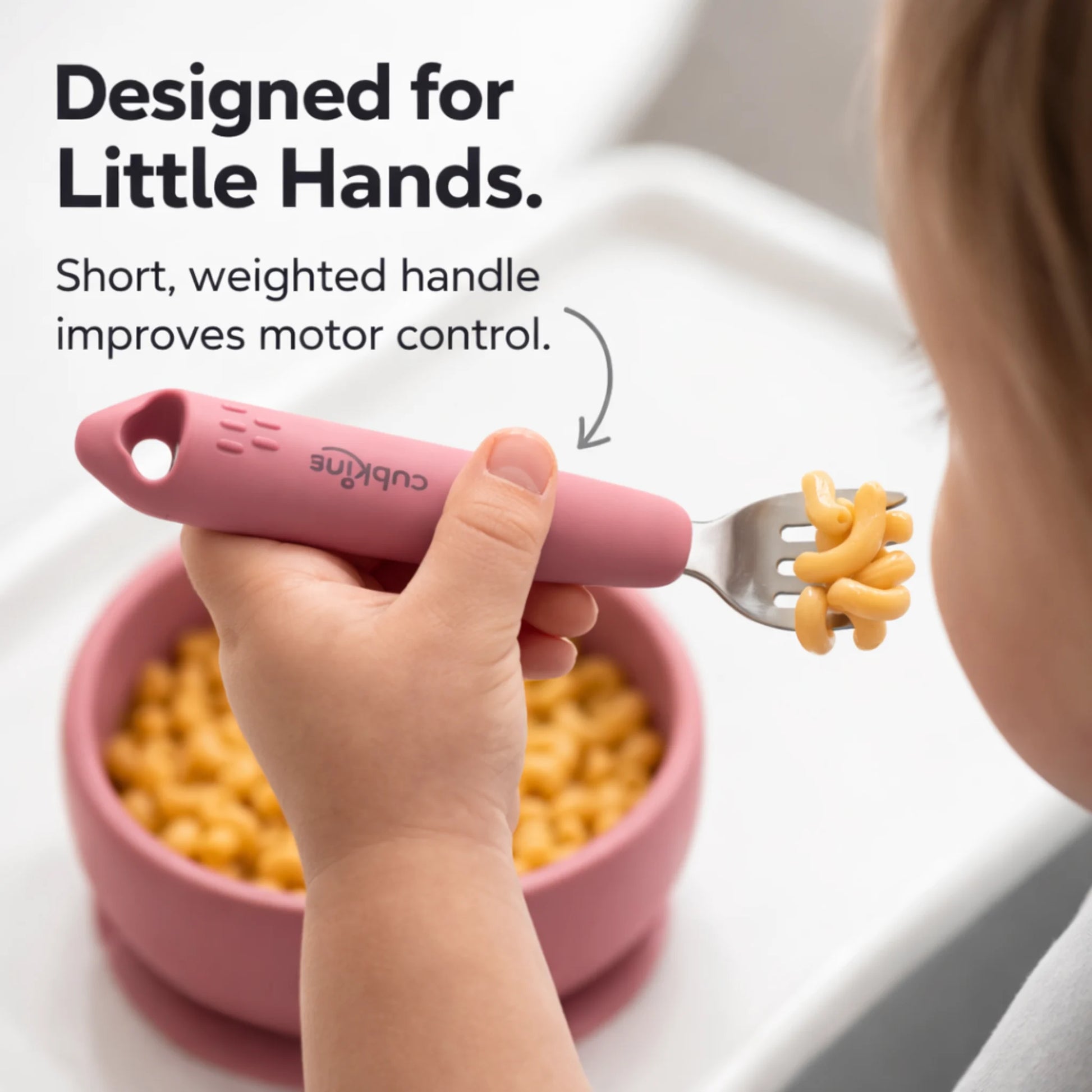 Toddler's hand holding a Cubkins Raspberry fork with macaroni on the tines above a matching raspberry silicone bowl, with the text "Designed for Little Hands – Short, weighted handle improves motor control"