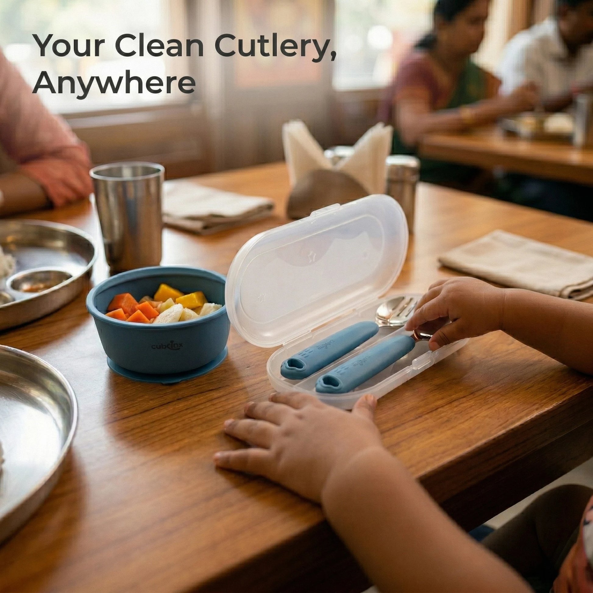Toddler reaching into an open Cubkins blue cutlery travel case at a restaurant table next to a silicone suction bowl with fruit