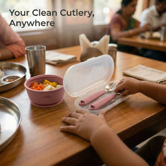 Toddler reaching into an open Cubkins raspberry cutlery travel case at a restaurant table next to a silicone suction bowl with fruit