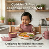 Baby wearing a raspberry Cubkins silicone bib self-feeding dal, rice and vegetables from a 4-compartment suction plate, with suction bowl, straw cup, steel spoon and fork on the table