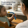 Indian baby from 6 months self-feeding with Cubkins blue bendable silicone spoon in a wooden high chair, with parent watching