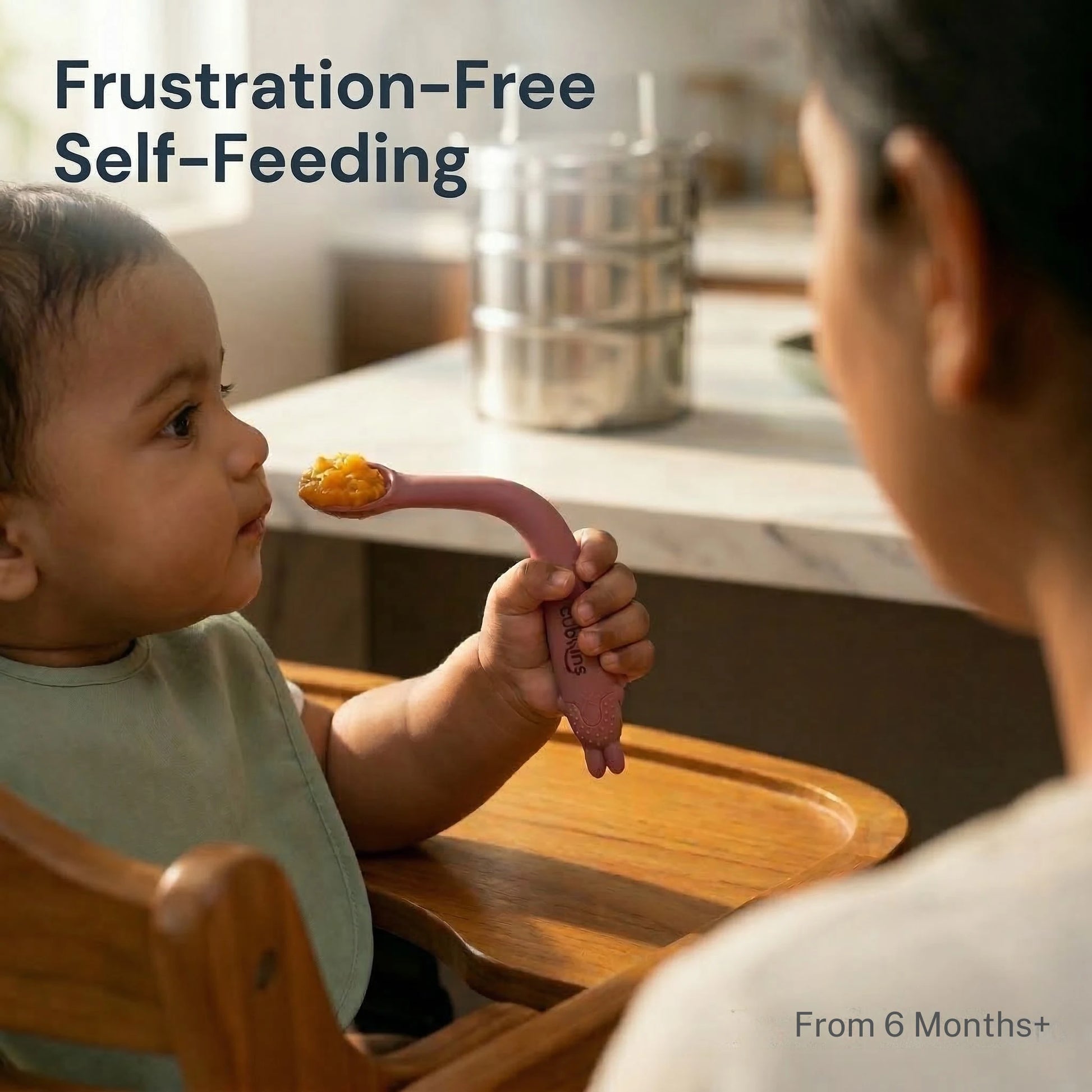 Indian baby from 6 months self-feeding with Cubkins raspberry pink bendable silicone spoon in a wooden high chair, with parent watching