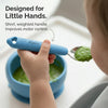 Baby's hand holding the short, weighted Cubkins spoon with a blue silicone handle, self-feeding green puree from a blue bowl.