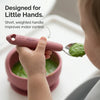 Baby's hand holding the short, weighted Cubkins spoon with a pink silicone handle, self-feeding green puree from a blue bowl.