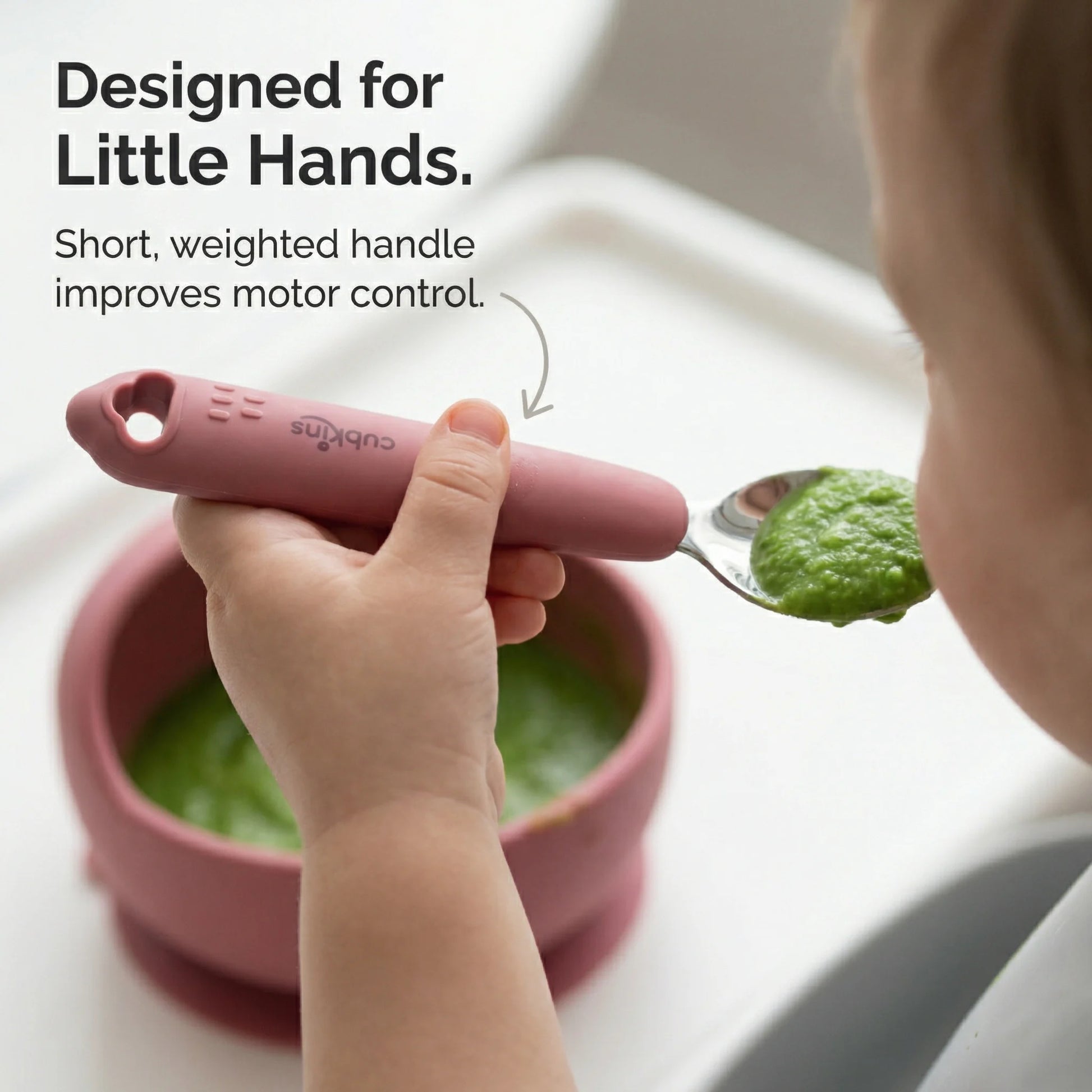 Baby's hand holding the short, weighted Cubkins spoon with a pink silicone handle, self-feeding green puree from a blue bowl.