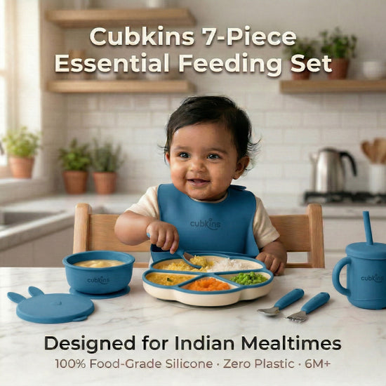 Smiling baby in a blue Cubkins silicone bib sitting at a marble kitchen counter with the full 7-piece Essential Feeding Set in blue — divided suction plate, suction bowl with bunny lid, straw sippy cup, and stainless steel cutlery — filled with dal, rice, and vegetables.