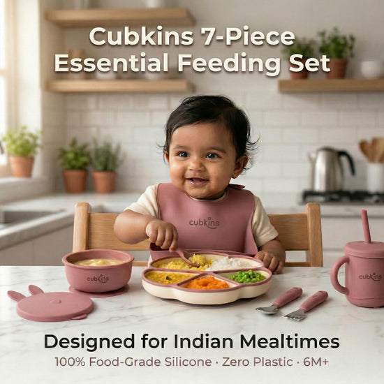 Smiling baby in a raspberry Cubkins silicone bib sitting at a marble kitchen counter with the full 7-piece Essential Feeding Set in raspberry — divided suction plate, suction bowl with bunny lid, straw sippy cup, and stainless steel cutlery — filled with dal, rice, and vegetables.