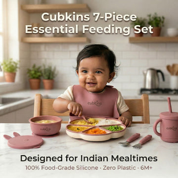 Smiling baby in a raspberry Cubkins silicone bib sitting at a marble kitchen counter with the full 7-piece Essential Feeding Set in raspberry — divided suction plate, suction bowl with bunny lid, straw sippy cup, and stainless steel cutlery — filled with dal, rice, and vegetables.