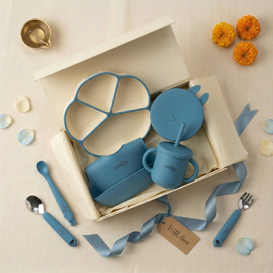 Overhead flatlay of the Cubkins blue 7-piece Essential Feeding Set presented as an Annaprashan gift — open cream gift box containing the divided suction plate, suction bowl with bunny lid, bib, and straw sippy cup, surrounded by scattered blue and white flower petals, a brass diya, orange marigold flowers, and a "With love" gift tag.