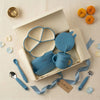 Overhead flatlay of the Cubkins blue 7-piece Essential Feeding Set presented as an Annaprashan gift — open cream gift box containing the divided suction plate, suction bowl with bunny lid, bib, and straw sippy cup, surrounded by scattered blue and white flower petals, a brass diya, orange marigold flowers, and a "With love" gift tag.