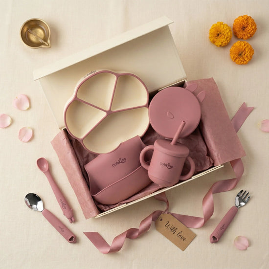 Overhead flatlay of the Cubkins raspberry 7-piece Essential Feeding Set presented as an Annaprashan gift — open cream gift box containing the divided suction plate, suction bowl with bunny lid, bib, and straw sippy cup, surrounded by scattered pink and white flower petals, a brass diya, orange marigold flowers, and a "With love" gift tag.
