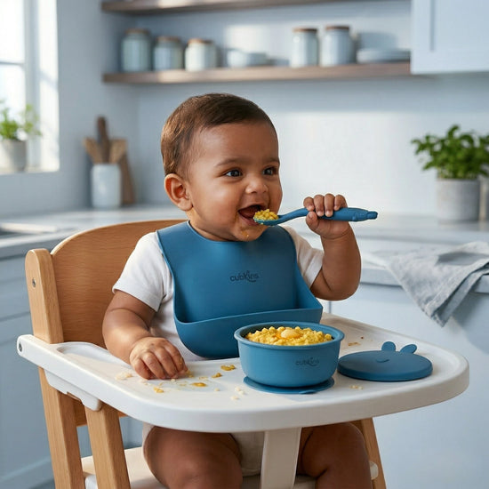 Indian baby boy eating from Cubkins blue silicone suction bowl using bendable spoon, wearing silicone bib in high chair