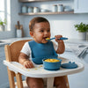 Indian baby boy eating from Cubkins blue silicone suction bowl using bendable spoon, wearing silicone bib in high chair
