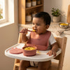 Indian baby eating from Cubkins raspberry silicone suction bowl using bendable spoon, wearing silicone bib in high chair