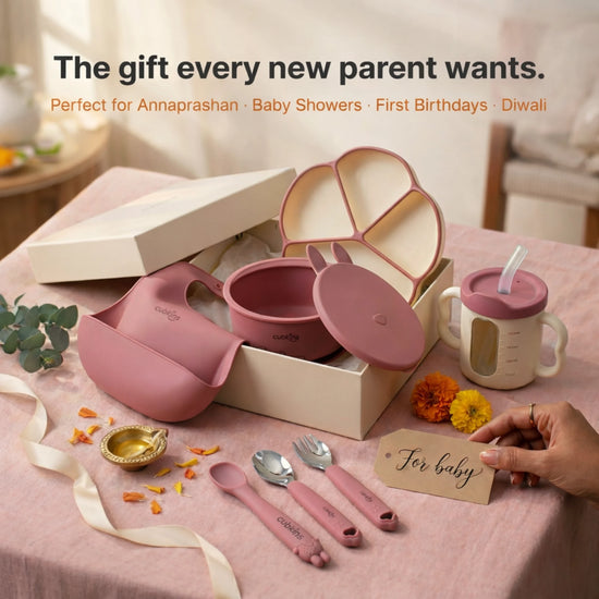 Cubkins 7-piece raspberry pink silicone baby feeding set presented as a gift in a premium cream box with a "For baby" tag, featuring suction plate, suction bowl with lid, bib, straw cup, spoon, fork and teether spoon — styled with marigolds and diya for Annaprashan, baby showers, first birthdays, and Diwali gifting