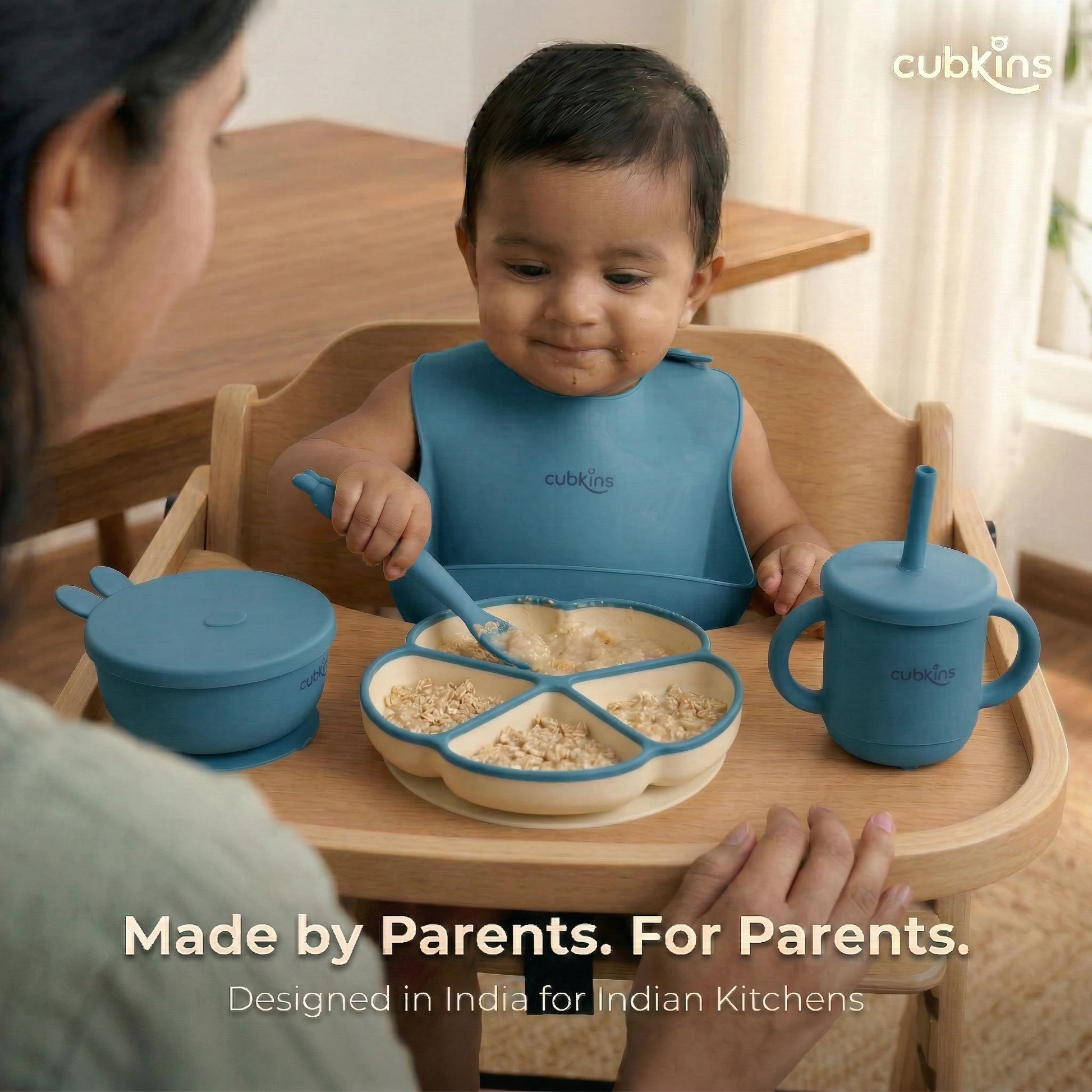 Indian mother watching over her baby seated in a wooden high chair, using the blue Cubkins 7-piece feeding set — divided suction plate filled with oats, suction bowl with bunny lid, and straw sippy cup. Text reads "Made by Parents. For Parents. Designed in India for Indian Kitchens.