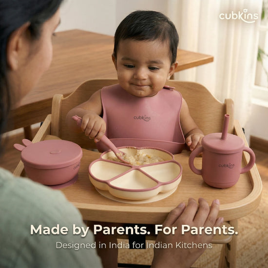 Indian mother watching over her baby seated in a wooden high chair, using the raspberry Cubkins 7-piece feeding set — divided suction plate filled with oats, suction bowl with bunny lid, and straw sippy cup. Text reads "Made by Parents. For Parents. Designed in India for Indian Kitchens.