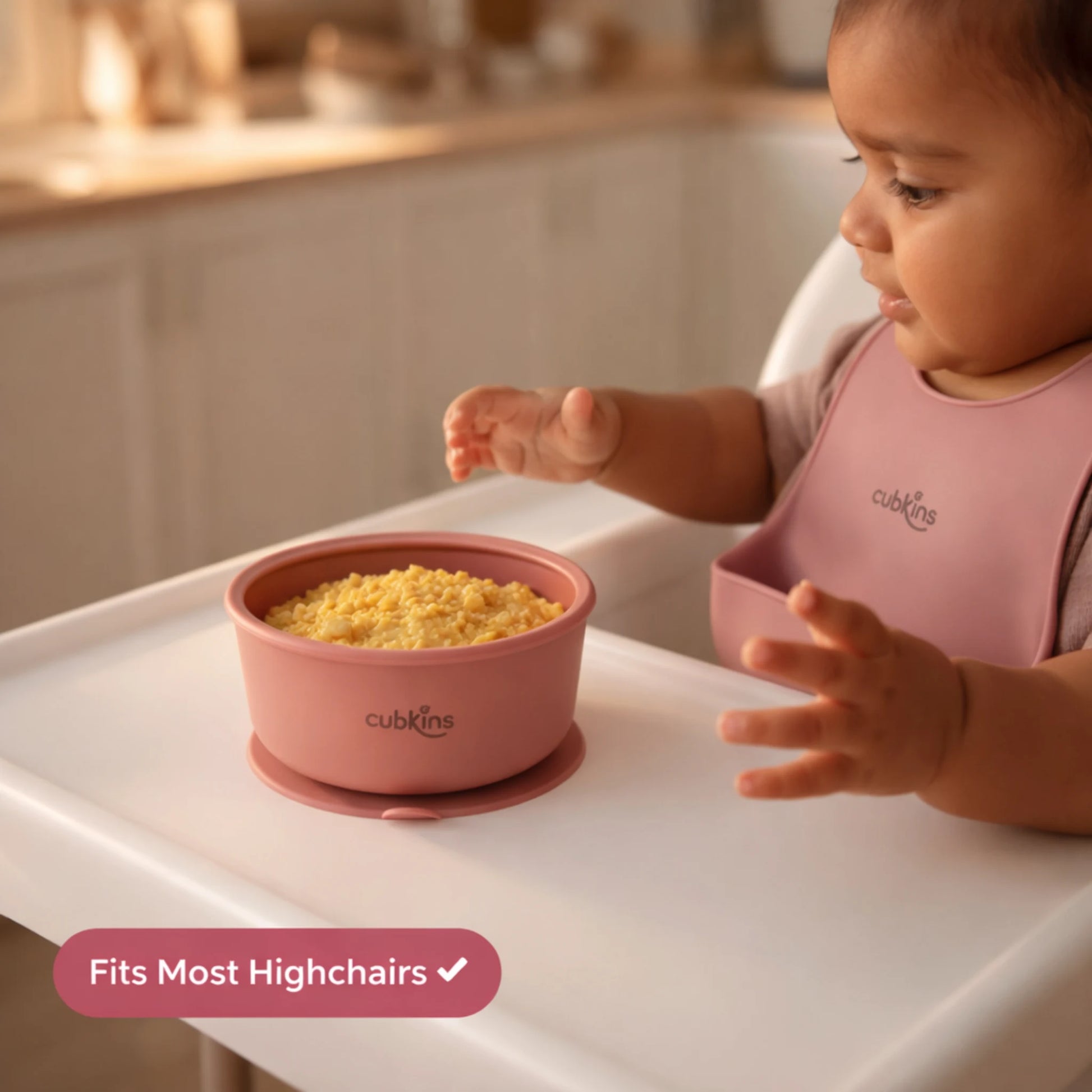Baby in pink bib eating from Cubkins raspberry silicone suction bowl on highchair tray showing fits most highchairs feature