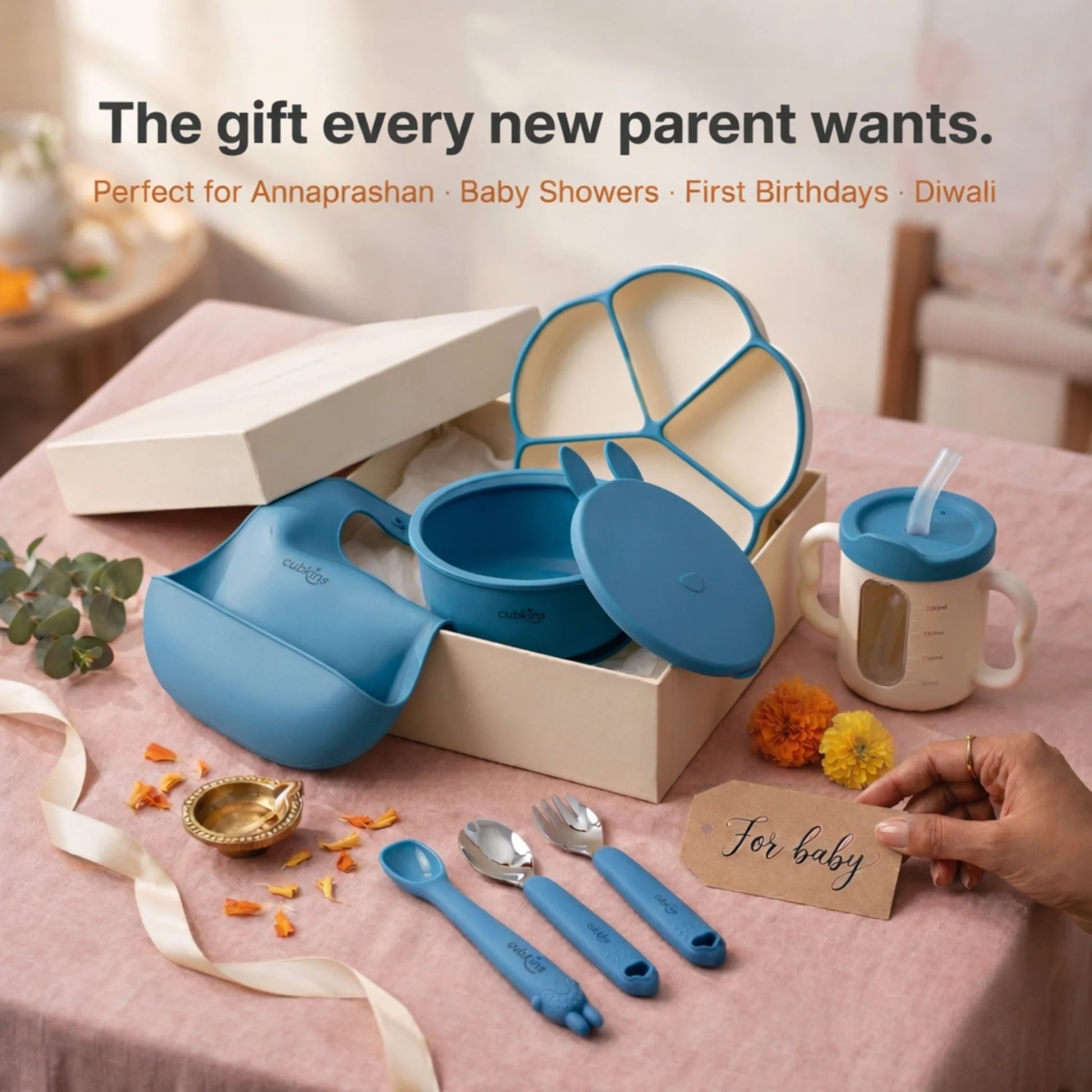 Cubkins 7-piece blue silicone baby feeding set presented as a gift in a premium cream box with a "For baby" tag, featuring suction plate, suction bowl with lid, bib, straw cup, spoon, fork and teether spoon — styled with marigolds and diya for Annaprashan, baby showers, first birthdays, and Diwali gifting
